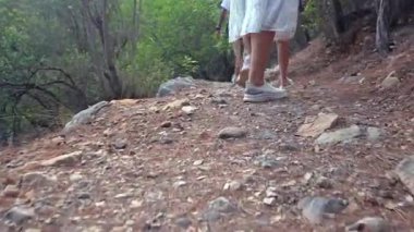 Bir grup yürüyüşçünün ormanda kuru ve kayalık bir yolda yürüdüğünü ve bacaklarına ve ayakkabılarına odaklandığını alçak açılı görüyor. 