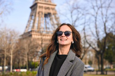 Güneşli bir bahar gününde Eyfel Kulesi 'nin önünde gülümseyen uzun saçlı, güneş gözlüklü ve gri ceketli çekici genç bir kadın.. 