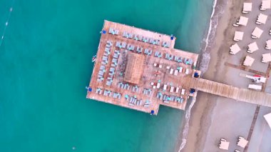 Yüksek açılı deniz manzaralı organize plaj şemsiyeleri, nazik kıyı dalgaları, ve kıyı şeridi yakınlarındaki bir rıhtımda turkuaz su. 