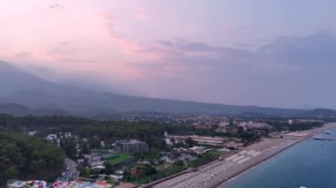 Gün batımında yoğun orman, uzak tepeler ve pastel renkli bulutlarla çevrili bir dağ kasabasının hava görüntüsü. 