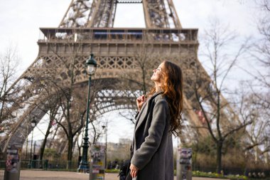 Paris 'te açık ve serin bir günde Eyfel Kulesi' nin yanında yürüyen ve gülümseyen uzun paltolu şık bir kadın.. 