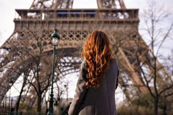 Gri paltolu, kendine güvenen kadın Eyfel Kulesi 'nin yanında duruyor doğal bir gülümsemesi ve rahat bir duruşu var.. 