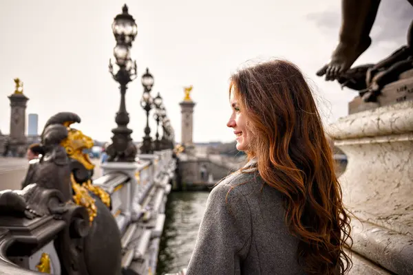 Uzun kıvırcık kumral saçlı ve gri paltolu kadın Paris 'teki süslü Alexander Köprüsü' nün üzerinde duruyor. Tarihi mimarinin ortasında Seine Nehri 'ne bakarken gülümsüyor.. 