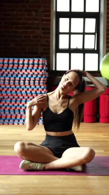 Genç bir kadın yoga minderinde bacak bacak üstüne atmış, başını yana eğerek güneşli bir spor salonunda, ahşap zeminli ve tuğladan duvarlı..