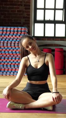 Genç bir kadın yoga minderinde bağdaş kurarak oturuyor. Ahşap duvarları ve ahşap duvarları olan parlak bir spor salonunda başını nazikçe yana eğiyor..