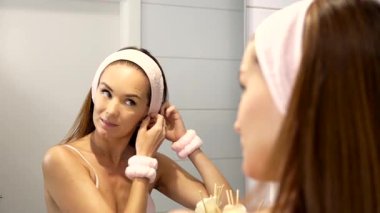 Genç bir kadın aynanın karşısında nazikçe saç bandını düzeltiyor. Günlük özgüven ve güzellik rutininin bir parçası olarak rahatlatıcı bir yüz bakımı seansına hazırlanıyor..
