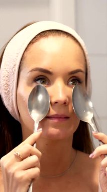 Evde serinletici bir yüz masajı yapmak için çene hizasına soğuk metal kaşıklar bastıran genç bir kadının dikey yakın çekimi. Bu cilt bakımı tekniği kaldırmayı ve kabartmayı destekler..