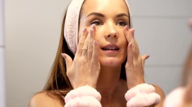 Genç bir kadın, avuçlarını yanaklarına bastırırken aynada kendine bakıyor. Bu jest, sabah yüz bakımı rutinini tamamlıyor. Huzurlu ve tazelenmiş bir cilt hissi bırakıyor..