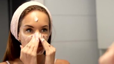 Banyodaki aynanın önünde yüzüne köpük temizleyici süren genç bir kadın. Saç bandı takarak cildini temizlemek ve tazelemek için günlük cilt bakımını yapar..