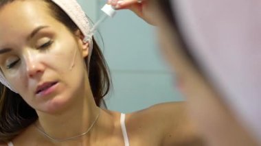 Genç bir kadın cam damlatıcısını kullanarak yüzüne günlük cilt bakımı uygulayarak sağlıklı ve ışıl ışıl bir cildi teşvik ediyor..