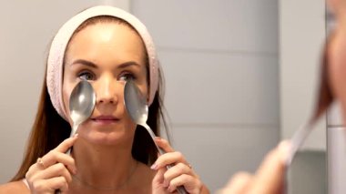 Genç bir kadın aynanın önünde duruyor ve şişkinliği azaltmak için soğuk metal kaşıkları nazikçe gözlerine bastırıyor. Bu doğal cilt bakımı, yüz rahatlığını ve göz altı bakımını destekler..