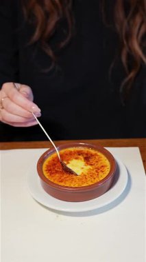 Kafe masasında oturan bir kadının yakın çekimi. Elinde kremalı Brulee tatlısının zengin muhallebisiyle dolu bir kaşık var. İçerideki sıcak ışığa yakalanmış..