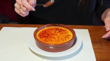 Beyaz bir tabakta kahverengi bir seramik tabakta servis edilen creme brulee tatlısının karamelize şeker kabuğunu kırmak için kaşık kullanan birinin yakın çekimi. Sıcak bir ortamda zengin doku.