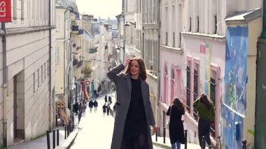 Paris, Fransa 04.01.2025 Gri montlu neşeli genç kadın Montmartre Paris 'te kaldırımlı bir caddede yürüyor, tarihi binalar ve arka planda yumuşak gün ışığı var.. 