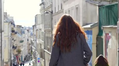 Paris, Fransa 04.01.2025 Dik kaldırımlı bir sokağa inen genç kadın. Parlayan güneş ışığı, klasik binalar ve sıradan yayalarla çevrili. Sadece editör kullanımı. 