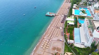 Şemsiyeli, havuzlu ve turkuaz Akdeniz kıyı şeridi boyunca ormanlık uçurumlu lüks bir sahil beldesinin yüksek açılı görüntüsü.