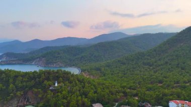 Yeşil tepeler ve yumuşak, renkli bir sabah gökyüzünün altındaki uzak sisli dağlarla çevrili gizli bir kıyı koyunun havadan görüntüsü. 