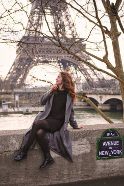 Paris 'te New York Bulvarı boyunca taştan bir duvarda oturan bir kadının görüntüsü, bulutlu bir günde arka planda Eyfel Kulesi ve Seine Nehri ile gülümsüyor. 