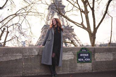 Paris 'te bulutlu bir günde arkasında Eyfel Kulesi olan gri ceketli şık bir kadın Seine nehrinin kenarında duruyor.