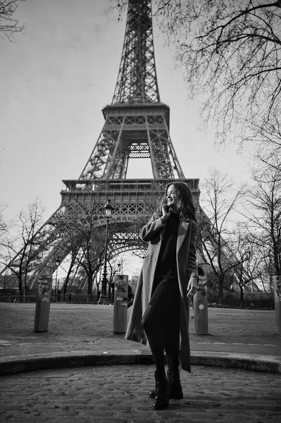 Paris 'te soğuk bir kış gününde Eyfel Kulesi' nin önünde gülümseyen zarif genç bir kadının siyah beyaz görüntüsü.