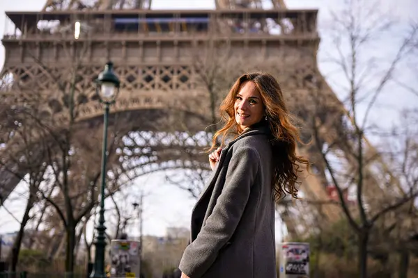 Eiffel Kulesi 'nin altında gülümseyen gri paltolu şık bir kadın. Etrafı çıplak ağaçlar ve karamsar gökyüzü ile çevrili.. 