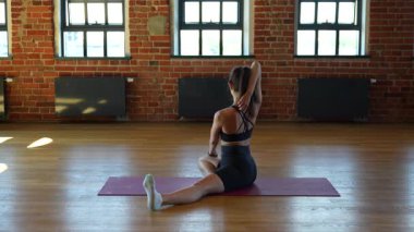 Geniş bacağının üzerinde, kolları başının arkasında eğilmiş, modern stüdyoda ahşap zemin ve tuğla duvarlarla yoga yapan genç bir kadının arka planı.