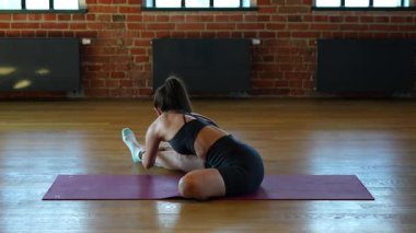 Bir bacağını öne eğmiş, sakin bir şekilde eğilmiş yoga minderinin üzerine yaslanmış tuğla duvarlar ve doğal ışıkla uzanmış genç bir kadının arka görüntüsü.