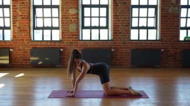 Aktif kıyafetli kadın dört ayak üstünde, inek pozu vermiş, arkası kemerli ve önüne bakıyor. Parlak çatı katında duvarları olan bir stüdyoda yoga yapıyor.