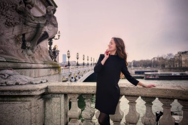 Paris 'te tarihi köprüde oturan şık genç bir kadın nehir manzarası ve arka planda klasik mimari. 