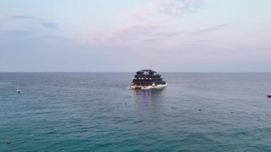 Sakin sularda seyreden modern bir çoklu güverte yolcu gemisinin insansız hava aracı görüntüleri yumuşak pastel renkli gökyüzünün altında sürekli yaklaşıyor. 