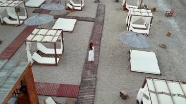 Yazlık otellerde, güneşli yataklarla kabinler arasında sahil kaldırımında yürüyen bir kadının hava manzarası. Altın saatte yakalanan, yaşam tarzı ve seyahat konsepti için insansız hava aracı görüntüleri.