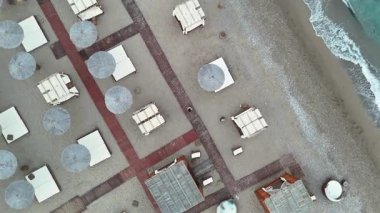 Sahil şeridi yakınında ahşap kaldırımlar, açık şemsiyeler ve kabin tarzı yapılar olan sakin bir sahil bölgesinin dönüşümlü hava manzarası. Bir insan, yukarıdan hafif bir ışığa yakalanmış, yürürken görülüyor..