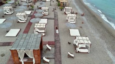 Deniz kıyısındaki oteldeki tahta tahta kaldırım boyunca kabinler ve şemsiyelerle yürüyen bir kadının insansız hava görüntüsü. Yaz boyunca kumlu sahil şeridinde yumuşak bir ışıkta yakalandılar..