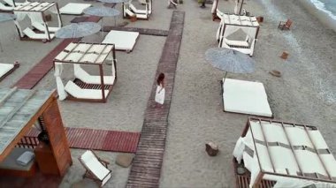 Beyaz elbiseli bir kadının, sessiz bir sahilde şemsiyeler ve şezlonglar arasındaki tahta patika boyunca yalınayak yürüdüğünü gösteren hava manzarası. Yumuşak gün ışığında yakalandı.