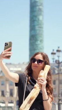 Genç bir kadın, Paris 'teki Place Vendome' da klasik mimariyle çevrili güneşli bir günde elinde baget tutarken akıllı telefonuyla gülümsüyor ve selfie çekiyor.