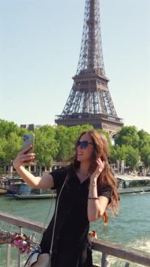 Gülümseyen genç kadın Paris 'in güneşli bir gününde Eiffel Kulesi' nin önünde romantik bir şehir köprüsünde aşk kilitleriyle duran akıllı telefonuyla selfie çeker.