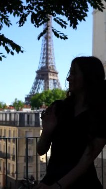 Paris 'te, ağaçlar ve yumuşak gölgelerle çerçevelenmiş güneşli bir günde arka planda Eyfel Kulesi ile güneş gözlüğünü ayarlayan genç bir kadının arka plandaki gölgesi.