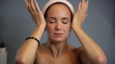 Kadın, yüz masajının son aşamasında avuçlarını alnına bastırıyor, gözleri kapalı ve rahat bir yüzle, evde özen ve farkındalık pratiği yapıyor.