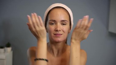 Yüzünde pembe bir saç bandı ve evde sakin bir ifade olan kadın yüz masajı rutininin bir parçası olarak iki avuç içi rahatlatıcı hareketle yüzünü kaplıyor.