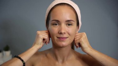 Gülümseyen kadın cilt bakımı sırasında eklemlerini kullanarak simetrik boyun masajı yapıyor. Pembe bir saç bandı takıyor ve rahat bir ev ortamında rahatlamış görünüyor.