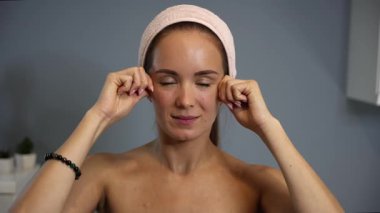 Kadın, yüz gerdirme rutininin bir parçası olarak yanaklarına masaj yapıyor. Sakin bir ev ortamında yukarı doğru hareket ettiğini ve cilt rengini gösteriyor.