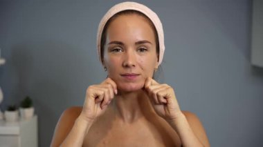 Kadın, yüz hareketleri sırasında çene kemiğine masaj yapıyor. Pembe saç bandı ve iç kısımda siyah bluz takarak kaldırma tekniği kullanıyor.