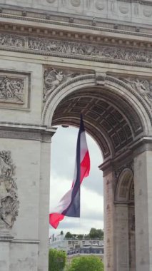 Paris, Fransa 'da Zafer Takı' nın altında rüzgarda dalgalanan Fransız üç renkli bayrağının dikey görüntüsü, detaylı taş mimarisi ve bulutlu gökyüzü..