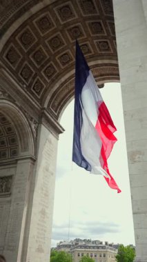 Arkasında bulutlu gökyüzü ve Haussmann mimarisi olan Paris 'teki Zafer Takı' nın altında dalgalanan Fransız ulusal bayrağının dikey görüntüsü