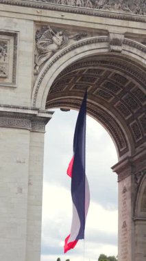 Fransa 'nın başkenti Paris' te Zafer Takı 'nın altında asılı Fransız üç renkli bayrağının dikey bileşimi, heykelsel detaylar ve arka planda bulutlu gökyüzü.