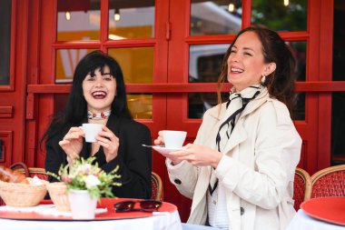 İki neşeli kadın, Paris 'teki bir kafede gülüyor ve kahve içiyor. Kırmızı sandalyeli ve çiçekli. Açık havada neşeli bir anı paylaşıyorlar..