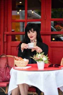 Zarif bir Paris kafesinde elinde bir fincan kahve tutan zarif bir kadın elinde kruvasan, çiçekler ve el çantasıyla kırmızı bir masada sessizce kameraya bakıyor..