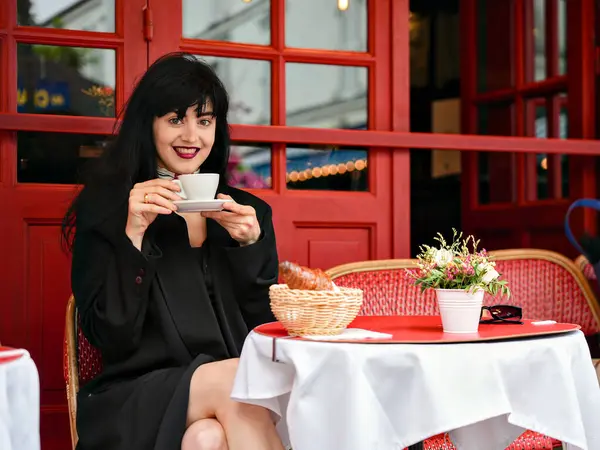 Neşeli bir kadın Paris 'te bir kafede oturuyor, gülümsüyor ve elinde bir fincan kahve, elinde kruvasan ve çiçeklerle klasik bir kırmızı arka planda oturuyor..