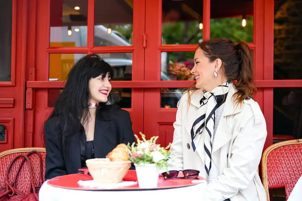 İki kadın Paris kafeteryasının terasında sohbet etmekten zevk alıyor. Kırmızı bir masada kruvasan ve kahveyle oturuyor, gülümsüyor ve açık havada samimi bir anı paylaşıyorlar..