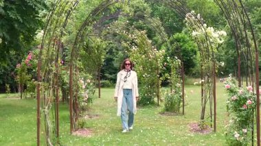 Parlak bir bahar gününde, dekoratif metal kemerlerin altında çiçek açan gül bahçesinde yürüyen yağmurluk ve güneş gözlüklü kendine güvenen bir kadın. Zarif ve taze mevsimlik açık hava manzarası.